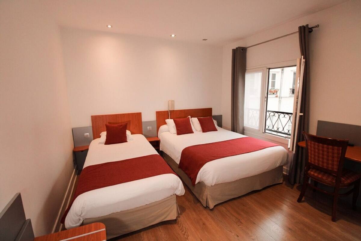 A hotel room with two beds, a wooden floor, a balcony with a railing, and a dining area with a chair and table.