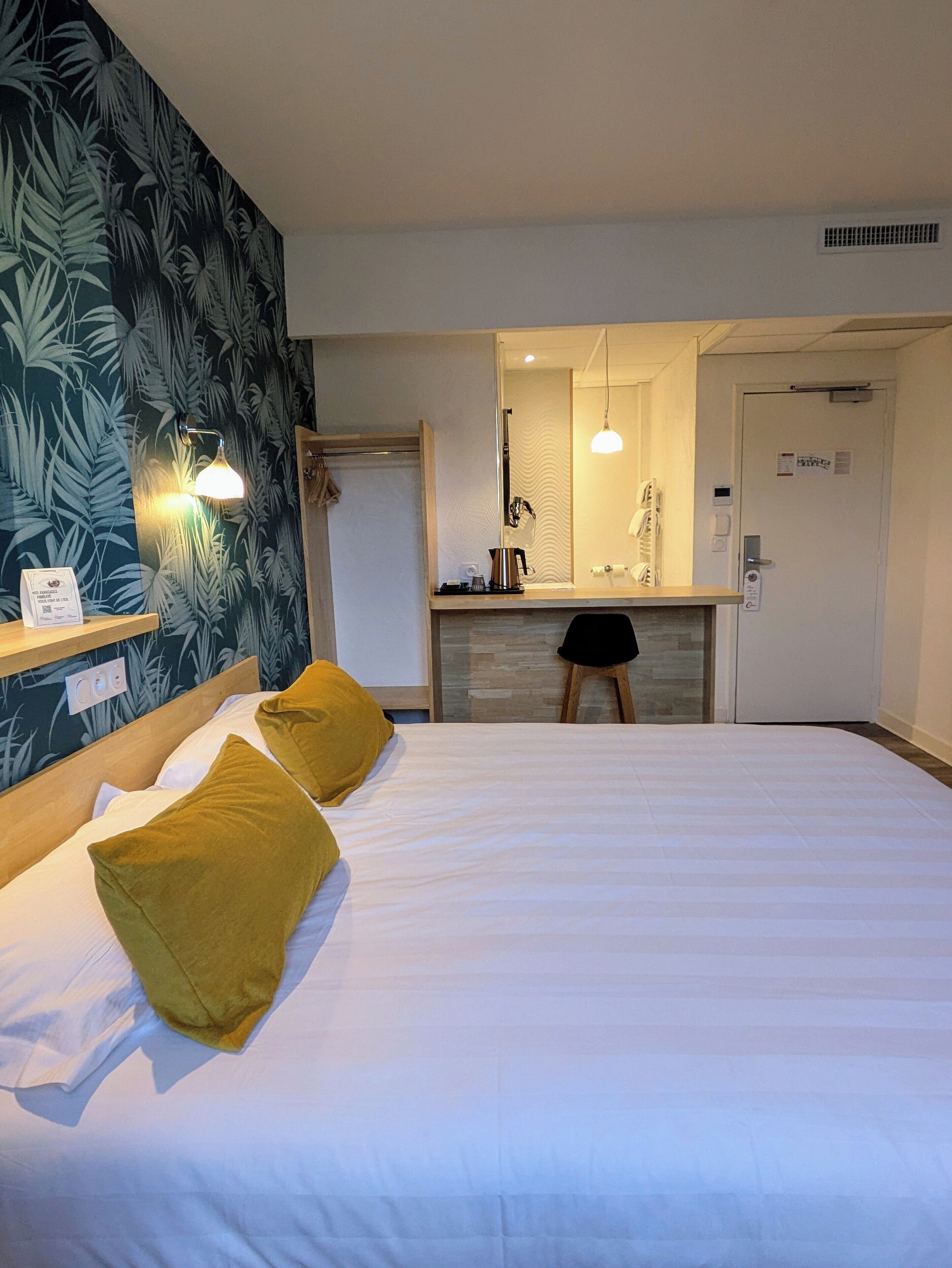Una habitación de hotel con cama, mesita de noche, espejo, lavabo y puerta.