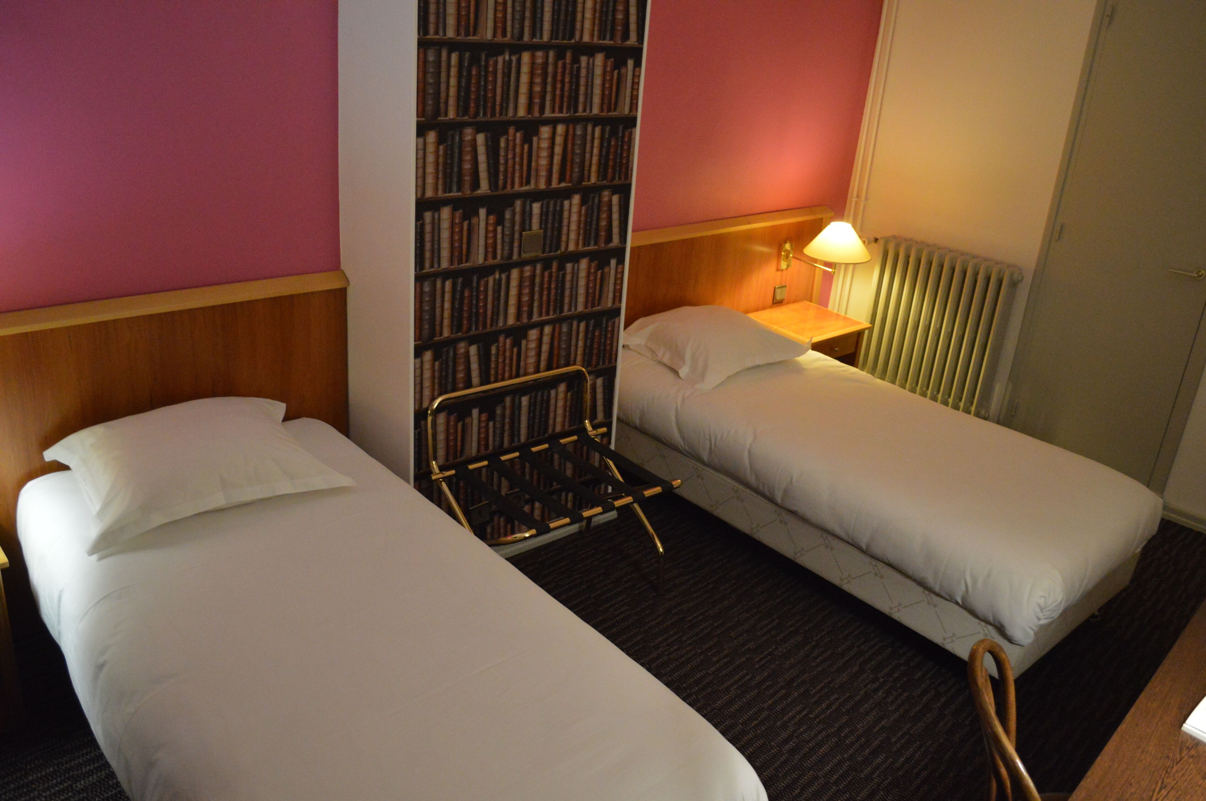A hotel room with two single beds, a bookshelf, a nightstand with a lamp, and a radiator.