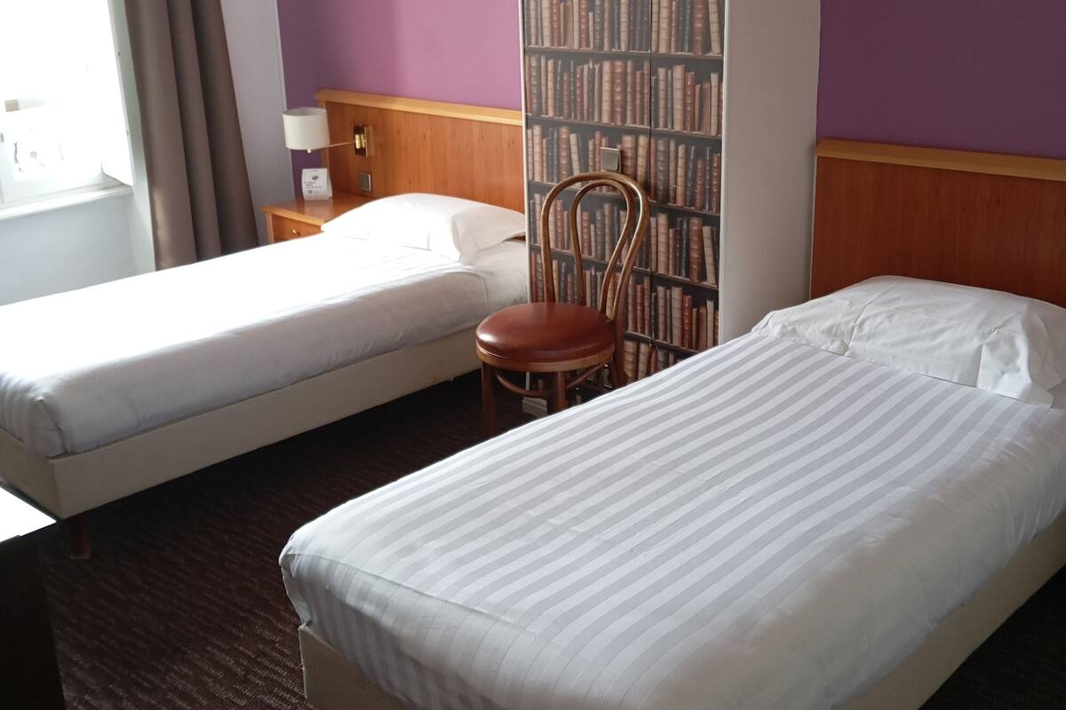 Ein Hotelzimmer mit zwei Einzelbetten, einem Holzstuhl, einem Bücherregal mit Büchern und einem Fenster mit Vorhängen.