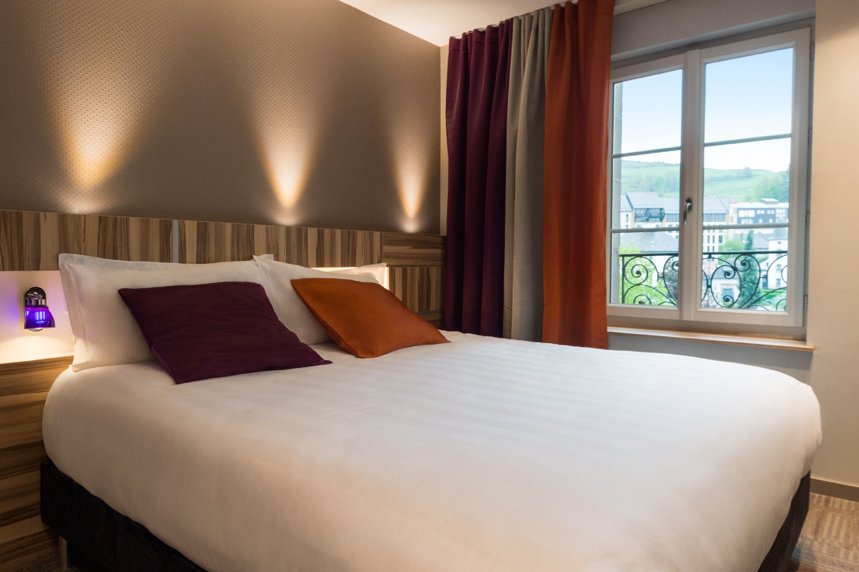 Una habitación de hotel con una cama grande, una mesita de noche con lámpara y una ventana con cortinas.