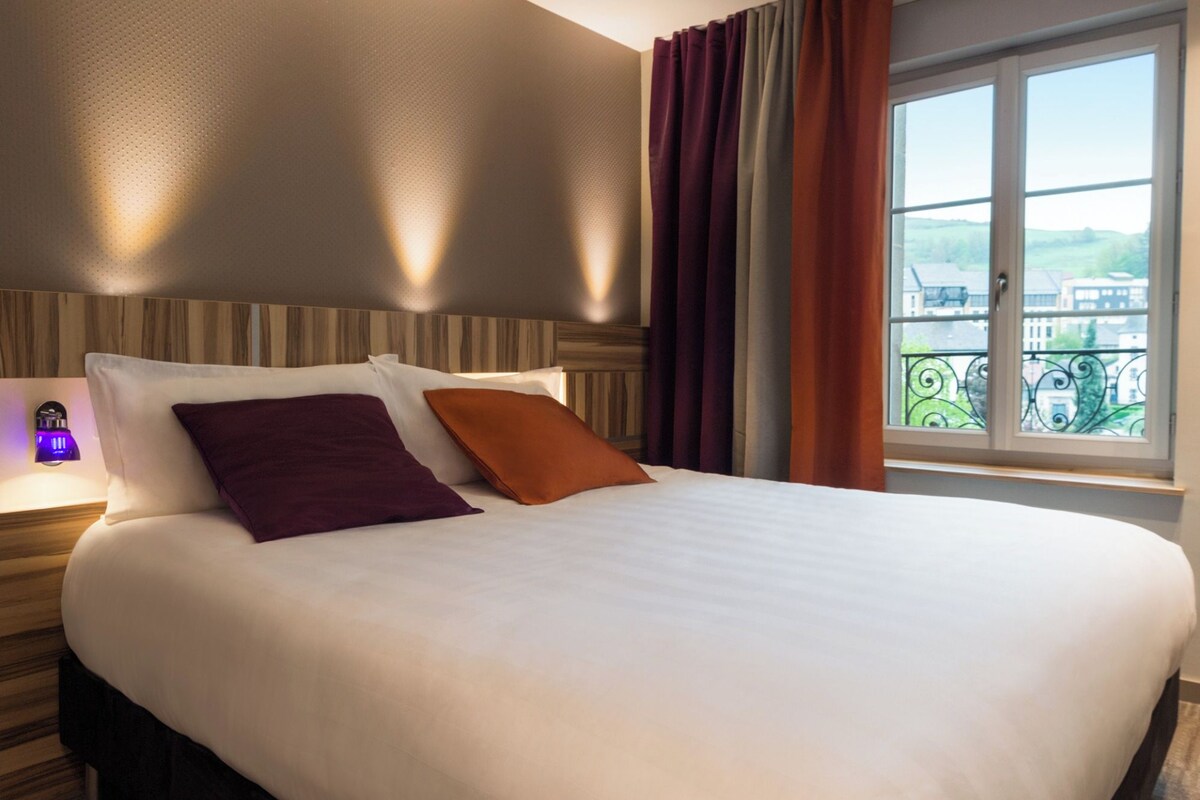 Habitación de hotel con una cama grande, una mesita de noche con lámpara y una ventana con vistas a edificios y zonas verdes.