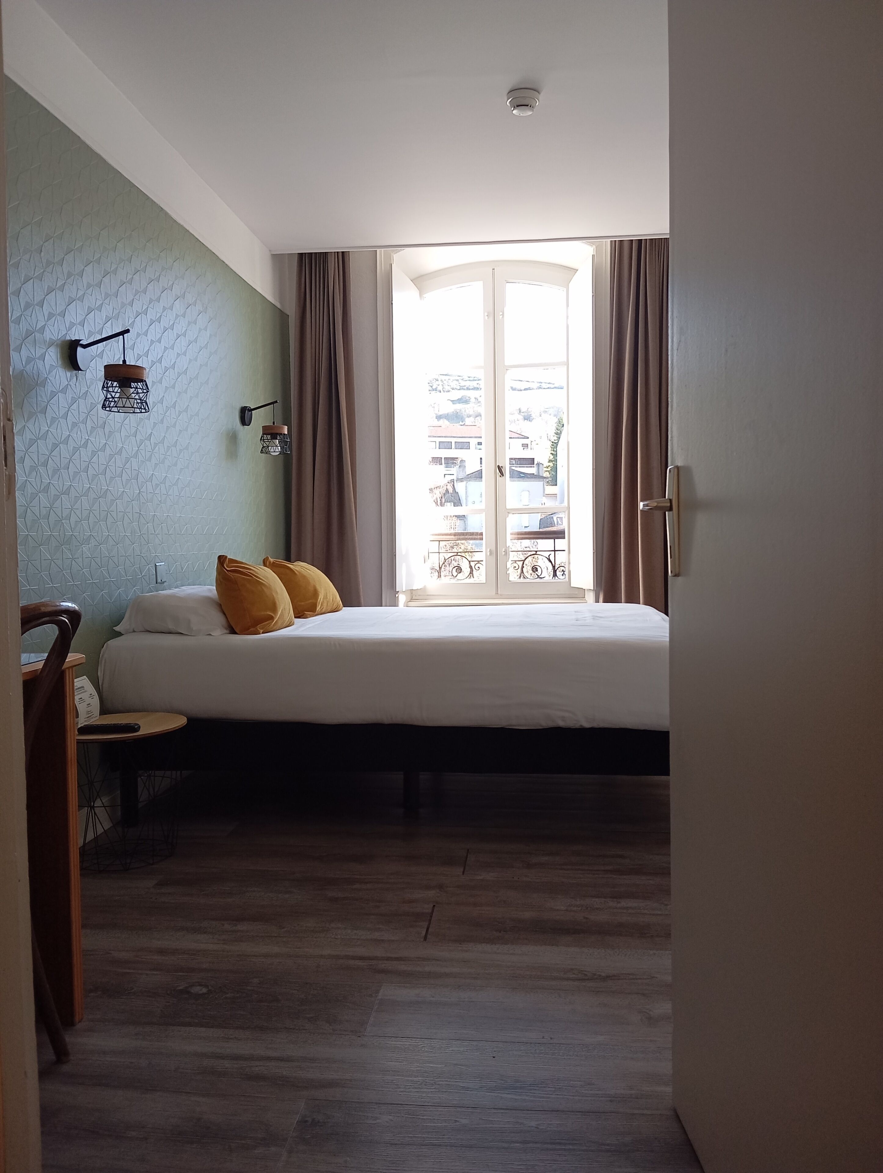 Un dormitorio con cama, una mesita, una silla, una ventana con cortinas y una lámpara fijada a la pared.