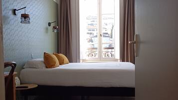 Un dormitorio con cama, una mesita, una silla, una ventana con cortinas y una lámpara fijada a la pared.
