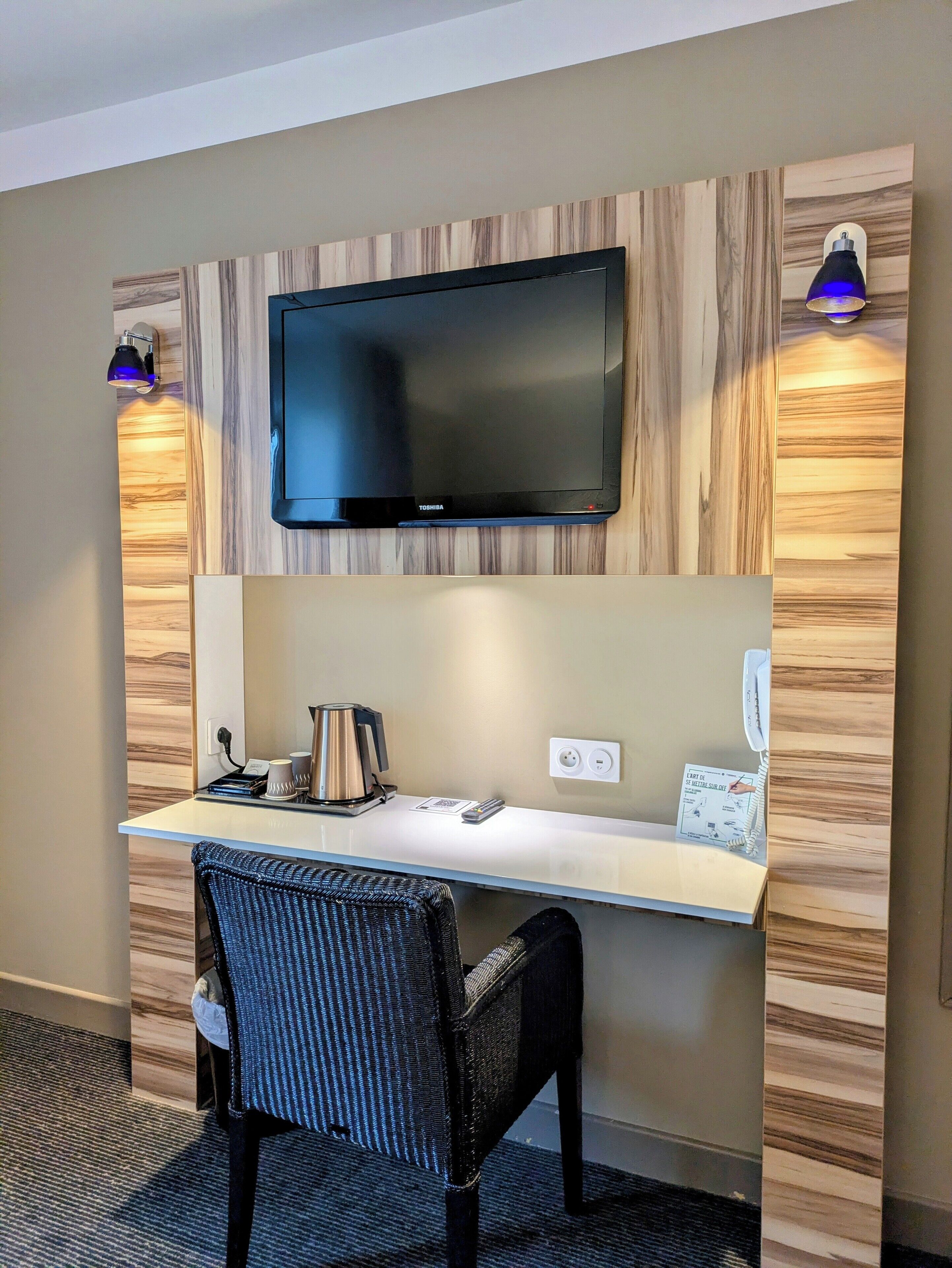 A hotel room with a flat-screen TV mounted on a wooden wall, a desk with a kettle, a chair, and a phone.