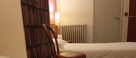 A hotel room with a single bed, a wooden chair, a bookshelf, a radiator, and a door.