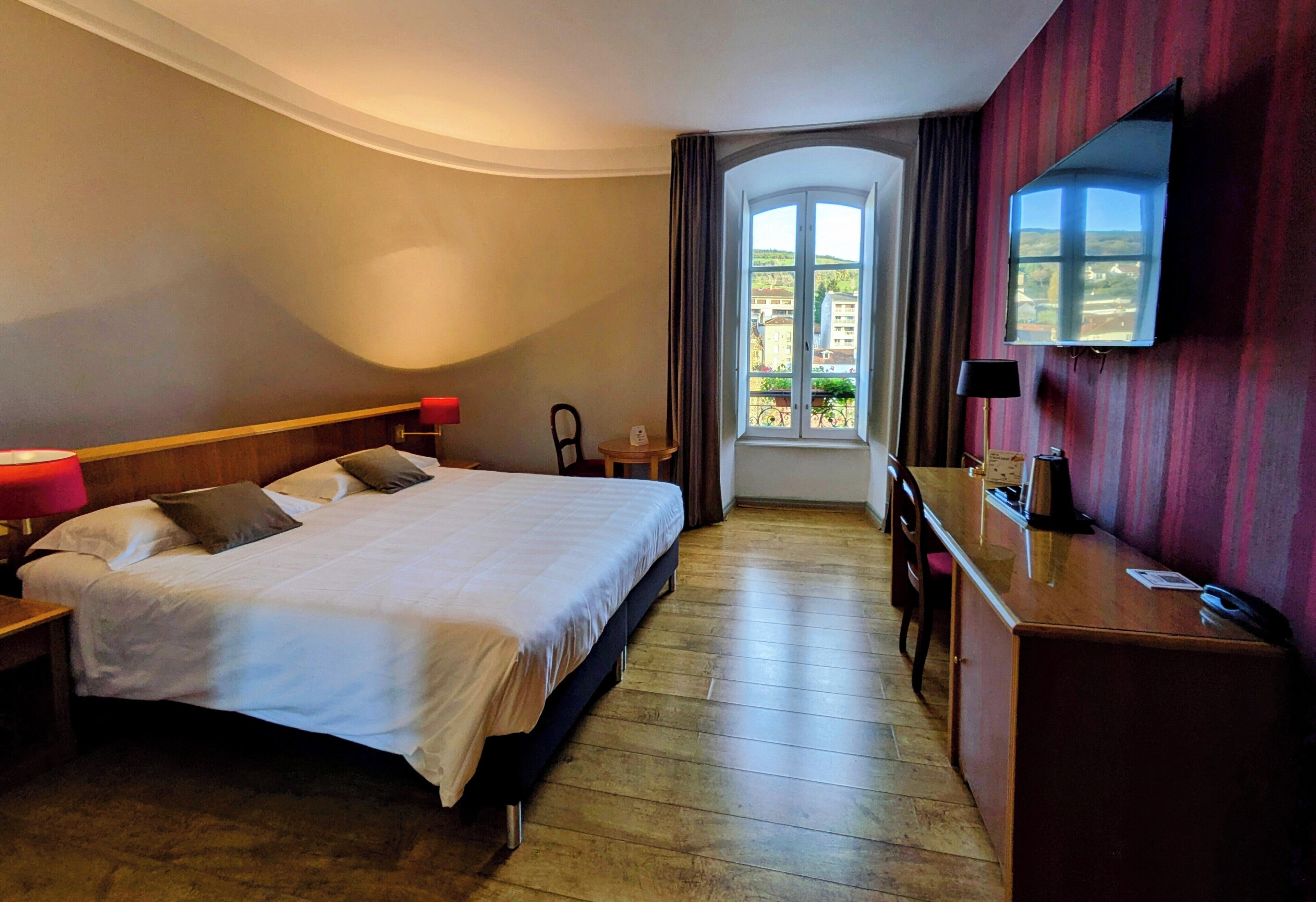 A hotel room with a large bed, a desk, a chair, a TV, and a window with a view of buildings.