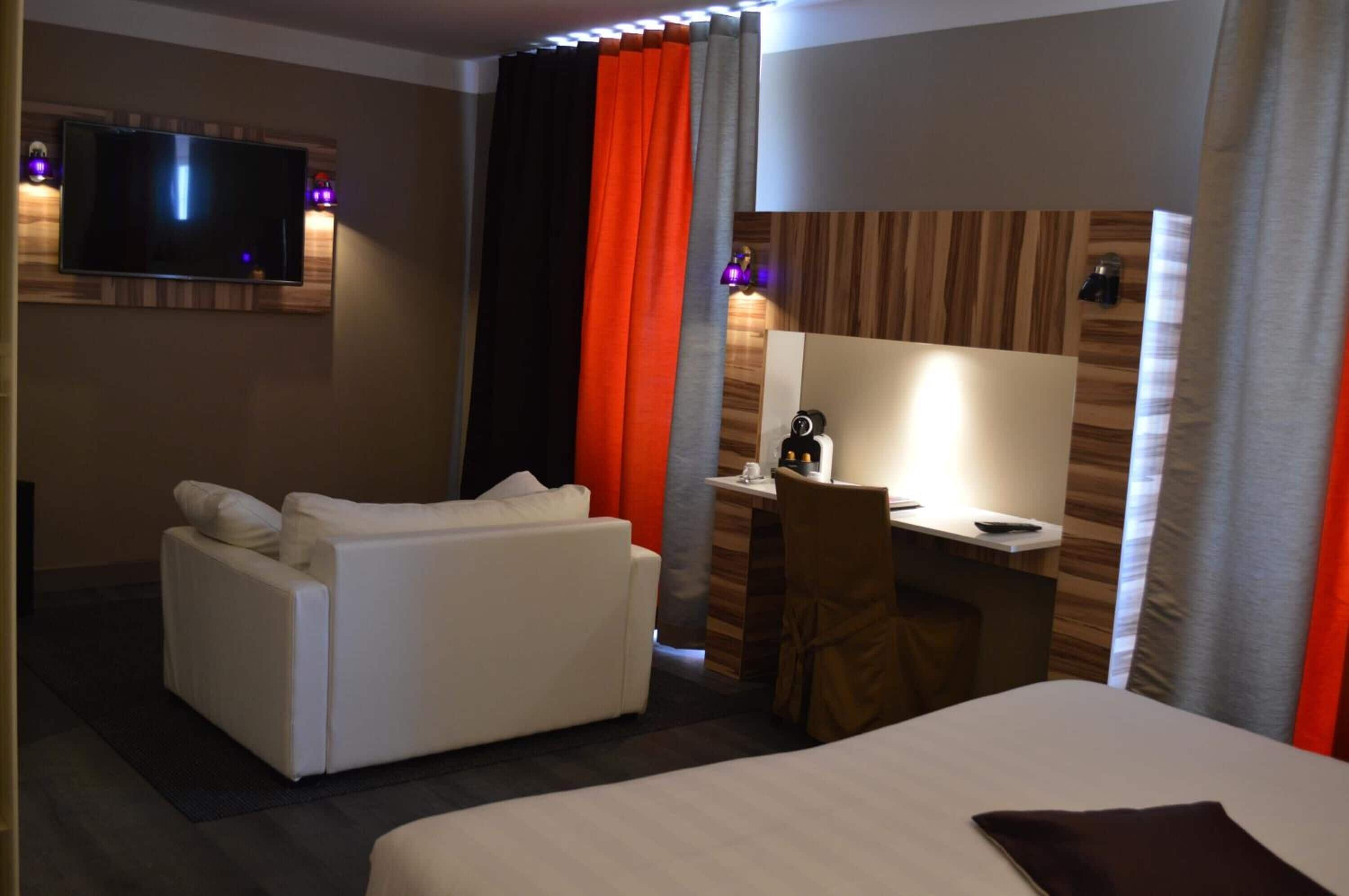 Hotelkamer met een bed, een slaapbank, een bureau met lamp, een aan de muur bevestigde televisie, en een gordijn met een rood en grijs patroon.