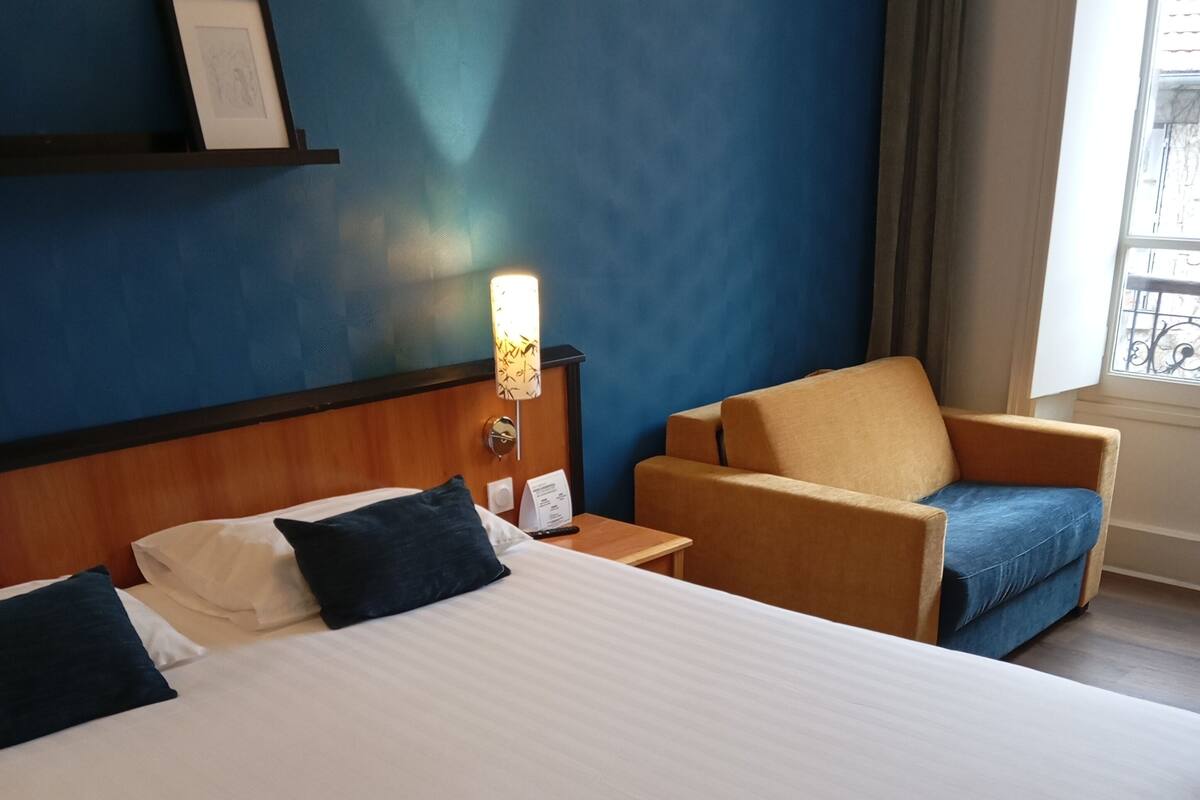 Ein Hotelzimmer mit einem großen Bett, einem Sessel, einem Nachttisch, einem an der Wand befestigten Regal mit einem Bild und einem Fenster mit Vorhängen.