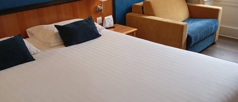 A hotel room with a large bed, a chair, a nightstand, a wall-mounted shelf with a picture, and a window with curtains.