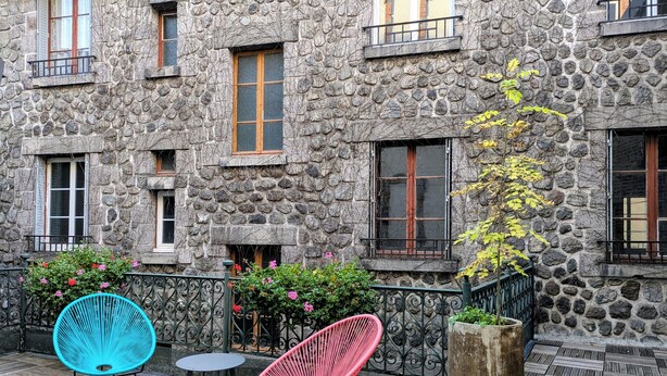 Un bâtiment en pierre doté de nombreuses fenêtres, un balcon avec une rambarde en métal et un espace extérieur aménagé avec deux chaises.