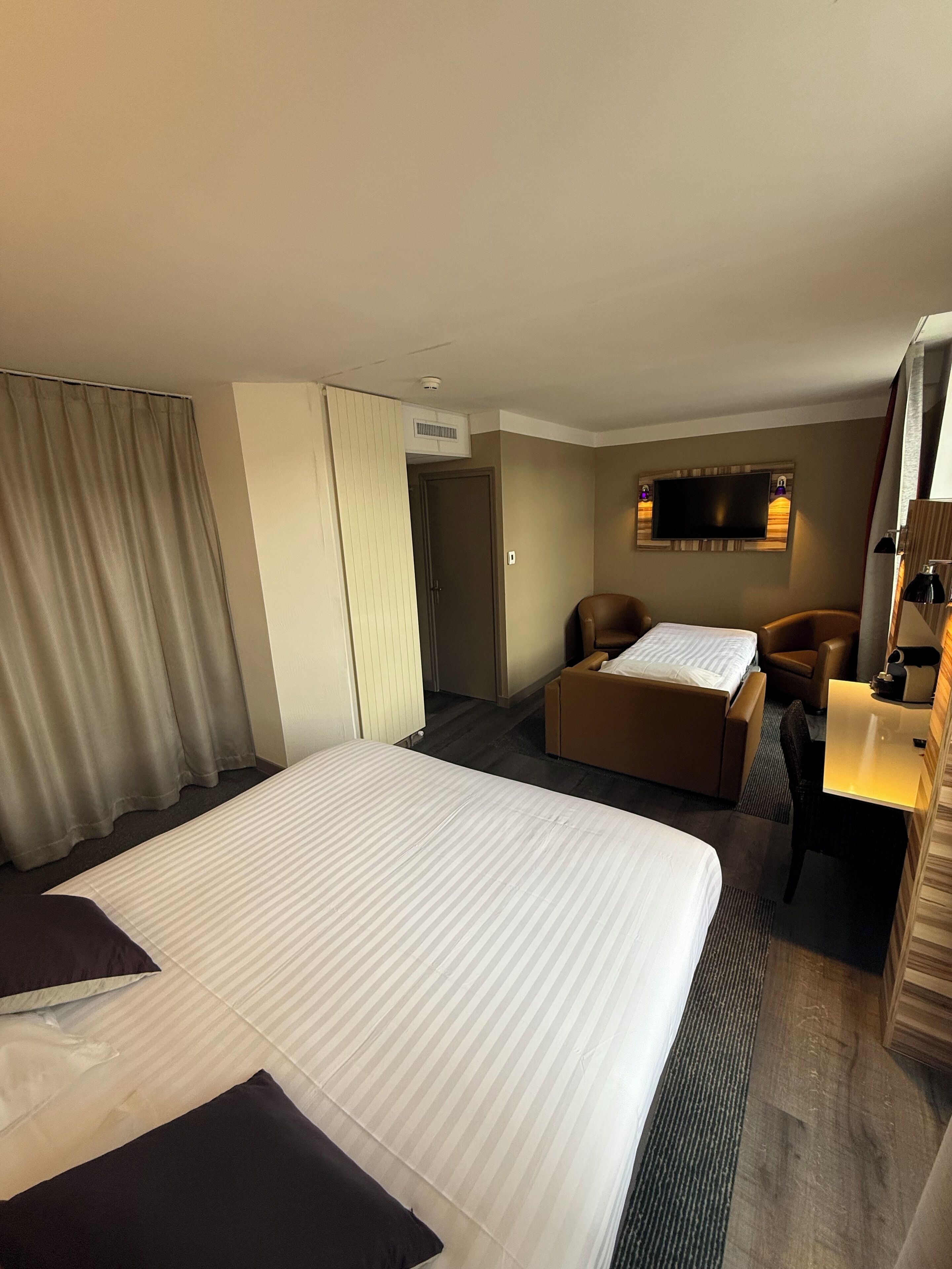 A hotel room with a large bed, a desk with a chair, a sofa, a television, and a window with curtains.