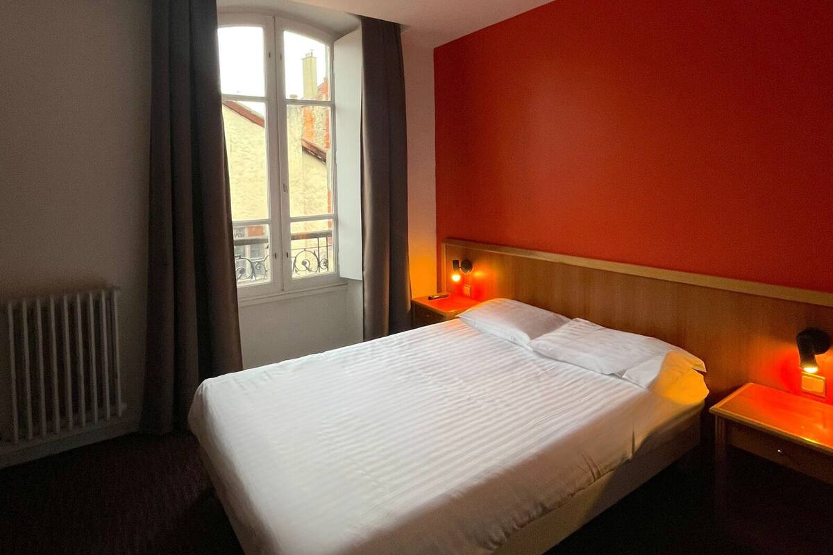 Ein Hotelzimmer mit einem großen Bett, einem Fenster mit Vorhängen, einem Heizkörper und zwei Nachttischlampen.