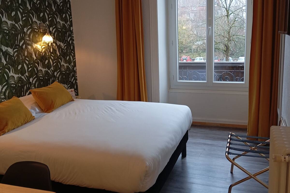 Ein Hotelzimmer mit einem Bett, einem Tisch, einem Stuhl, einem Fenster mit Vorhängen und einer Wand mit Blättermotiv.