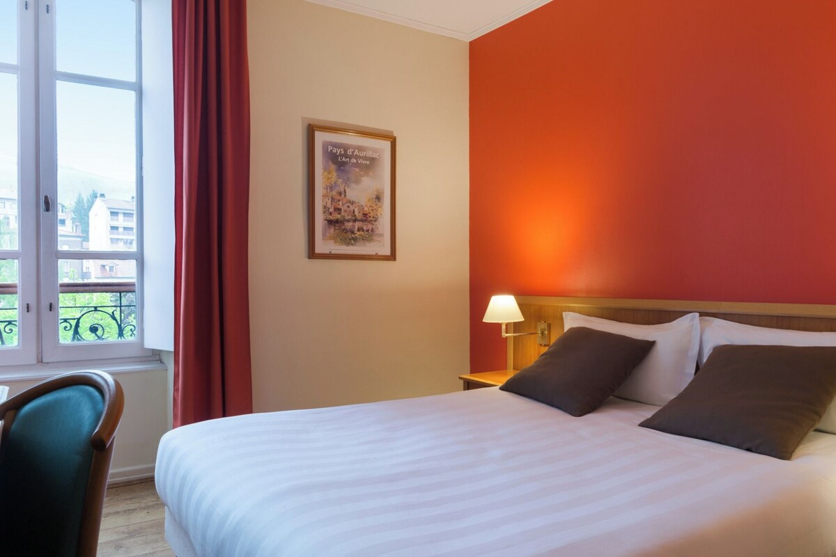 Una habitación de hotel con una cama, una mesita de noche, una lámpara, un cuadro enmarcado en la pared y una ventana con cortinas.