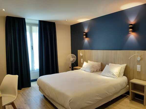 Una habitación de hotel moderna con una cama grande, dos mesitas de noche, un abanico de pie y una ventana con cortinas.