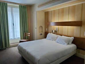 Habitación de hotel con una cama grande, cabecera de madera, una silla, una mesita de noche y una ventana con cortinas.
