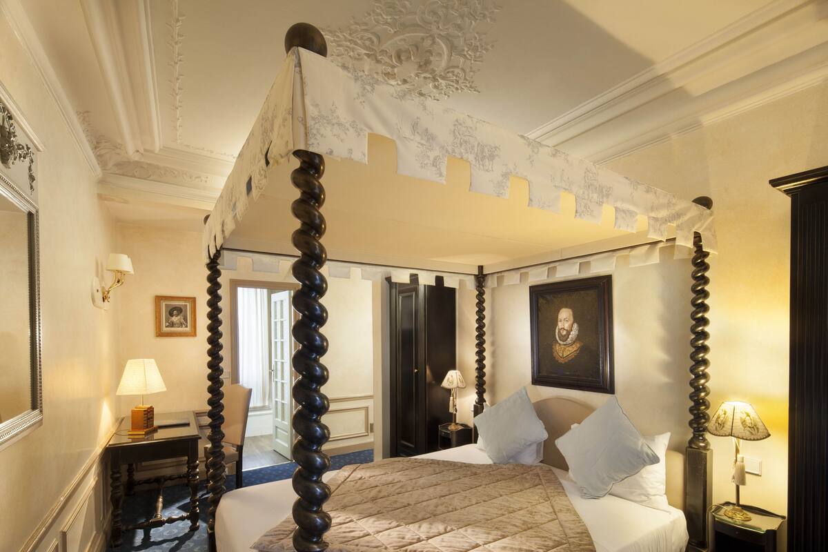A four-poster bed with a quilted bedspread, a nightstand with a lamp, and a framed portrait on the wall.