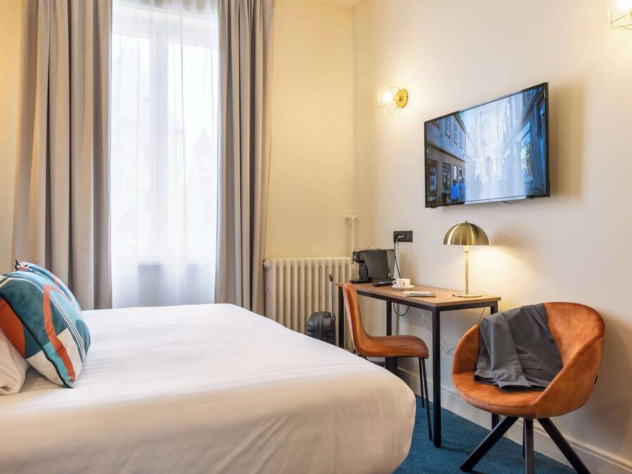 A hotel room with a bed, a desk with a computer, a chair, a lamp, and a painting on the wall.