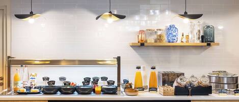 A breakfast bar with various food items and beverages, a shelf with decorative jars, and a stack of plates and bowls.