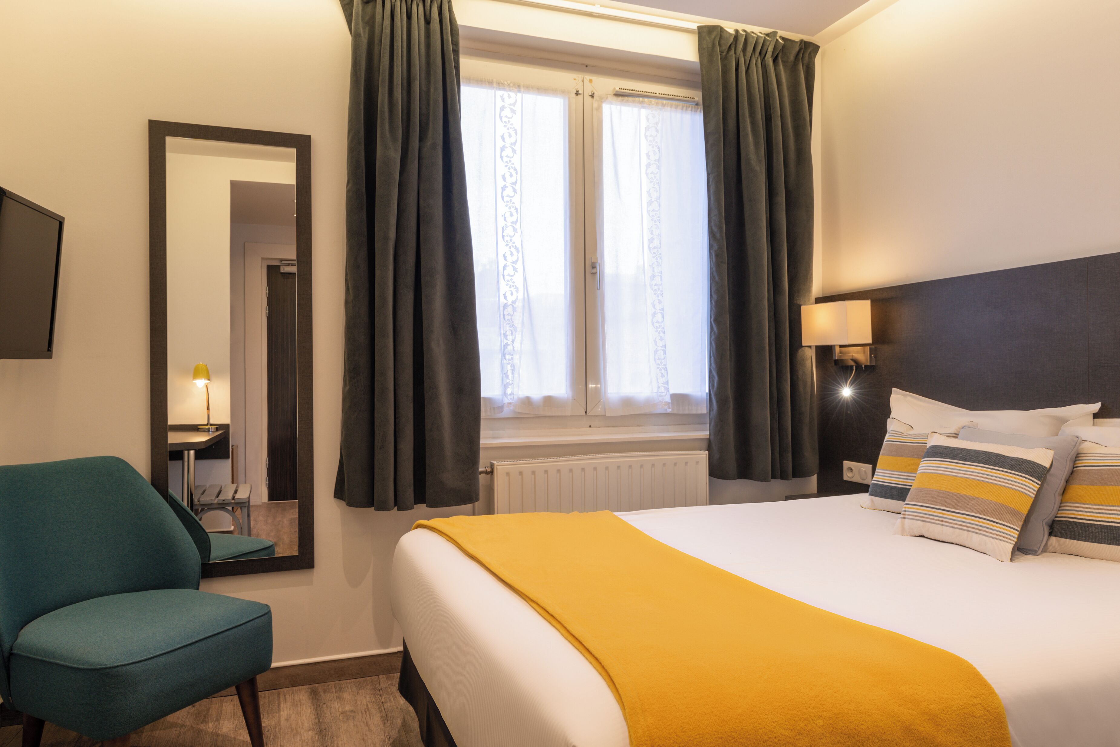 A hotel room with a bed, a chair, a mirror, a window with curtains, and a lamp.
