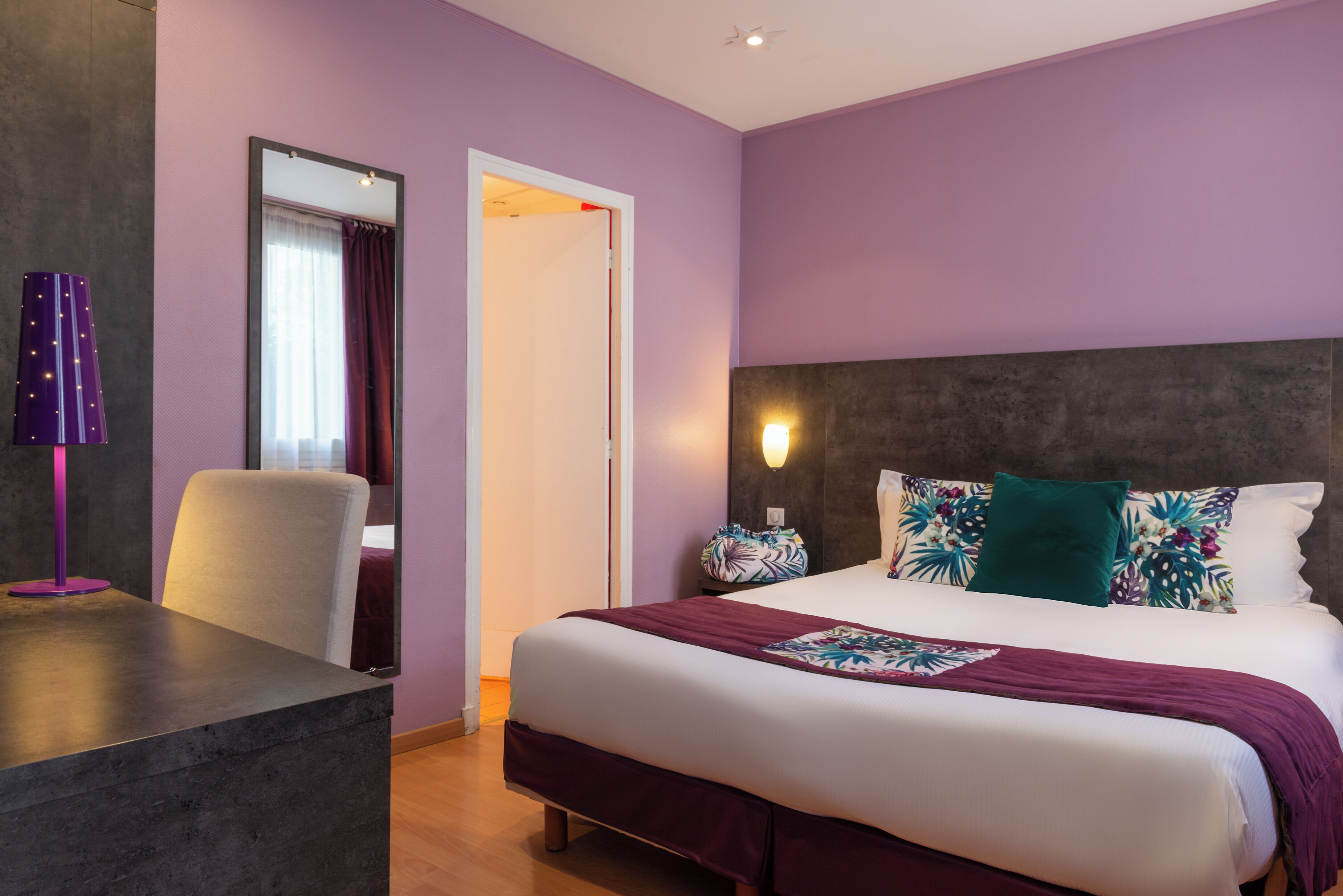 A hotel room with a large bed, a desk, a chair, a mirror, and a nightstand with a lamp.