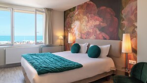 Habitación de hotel con una cama grande, dos lámparas de noche, vista al mar y un cuadro floral de gran tamaño en la pared.