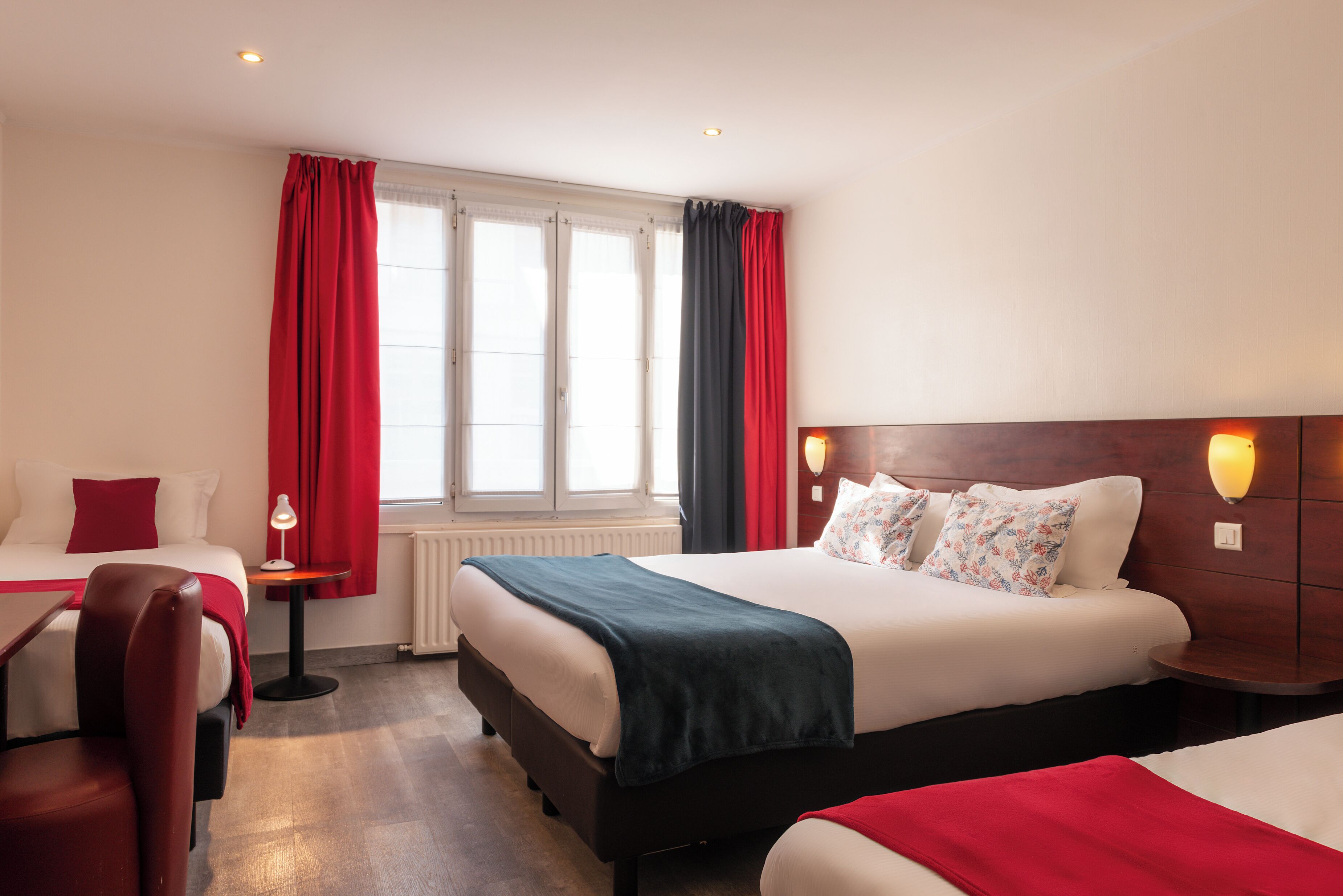 Une chambre d’hôtel avec deux lits, des rideaux rouges, une tête de lit en bois, une petite table avec une lampe et un radiateur.