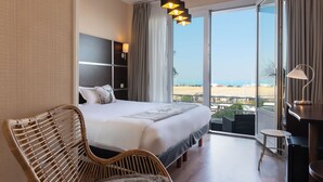 Habitación de hotel con una cama grande, una silla de mimbre, un escritorio y vista a la playa.