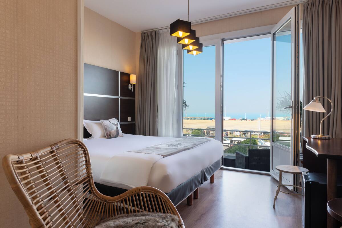 A hotel room with a large bed, a wicker chair, a desk, and a view of the beach.