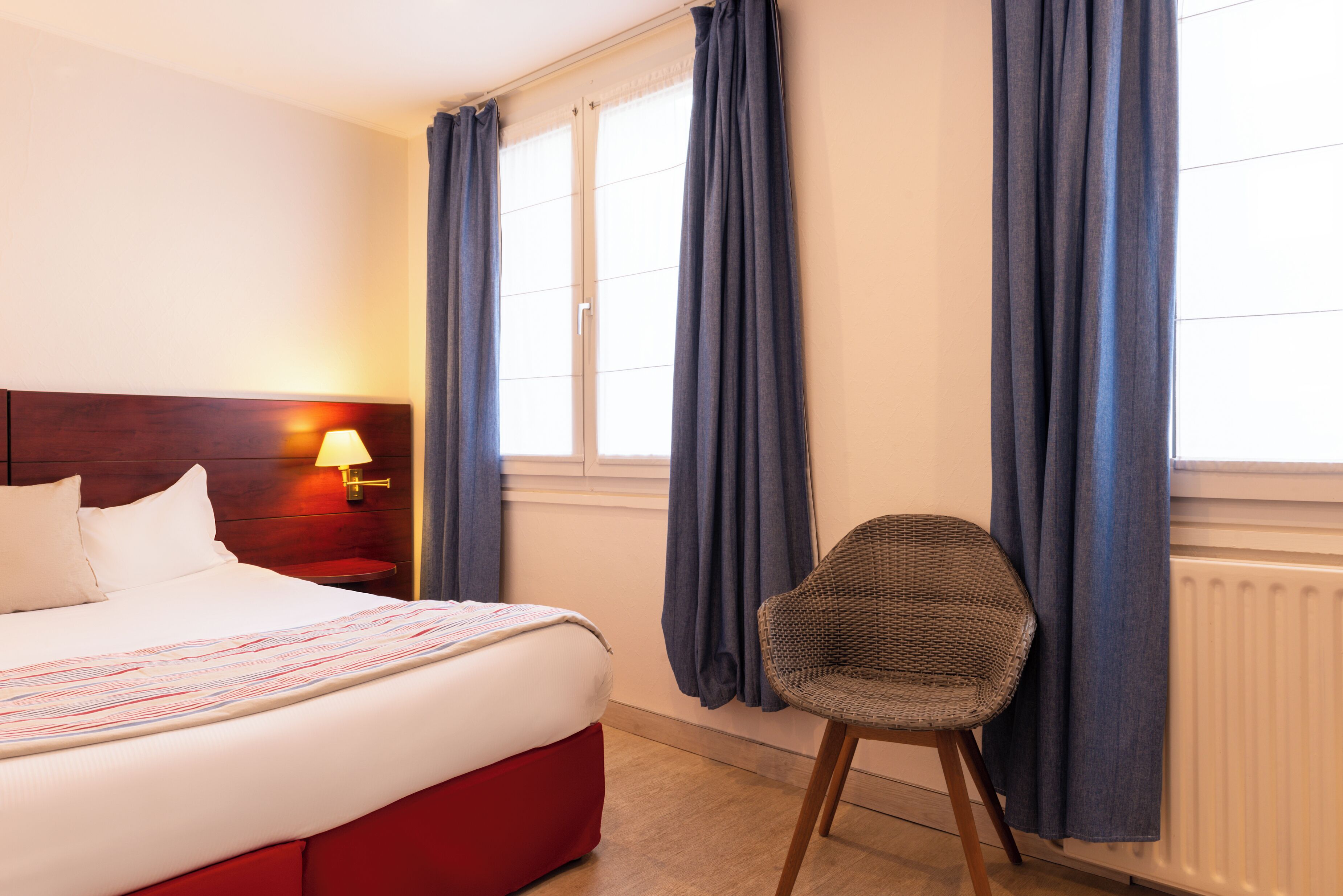 Una camera d'albergo con un letto, una sedia, un comodino con una lampada e una finestra con tende.
