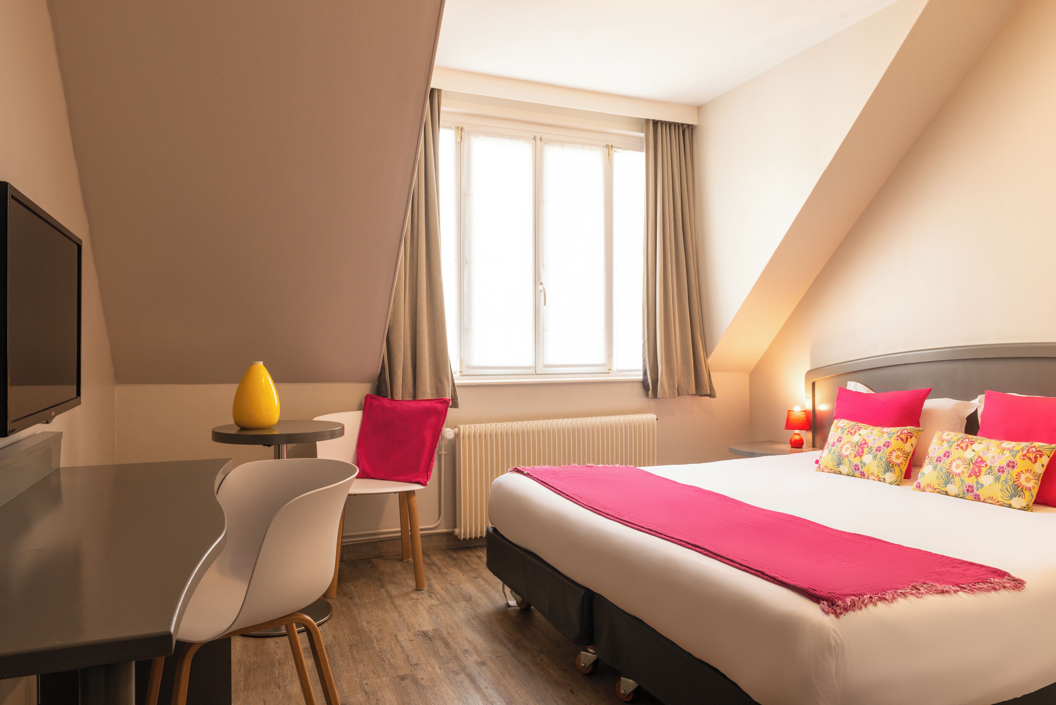Habitación de hotel con cama, escritorio, silla, mesita con un jarrón amarillo y ventana con cortinas.