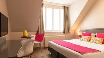 Habitación de hotel con cama, escritorio, silla, mesita con un jarrón amarillo y ventana con cortinas.