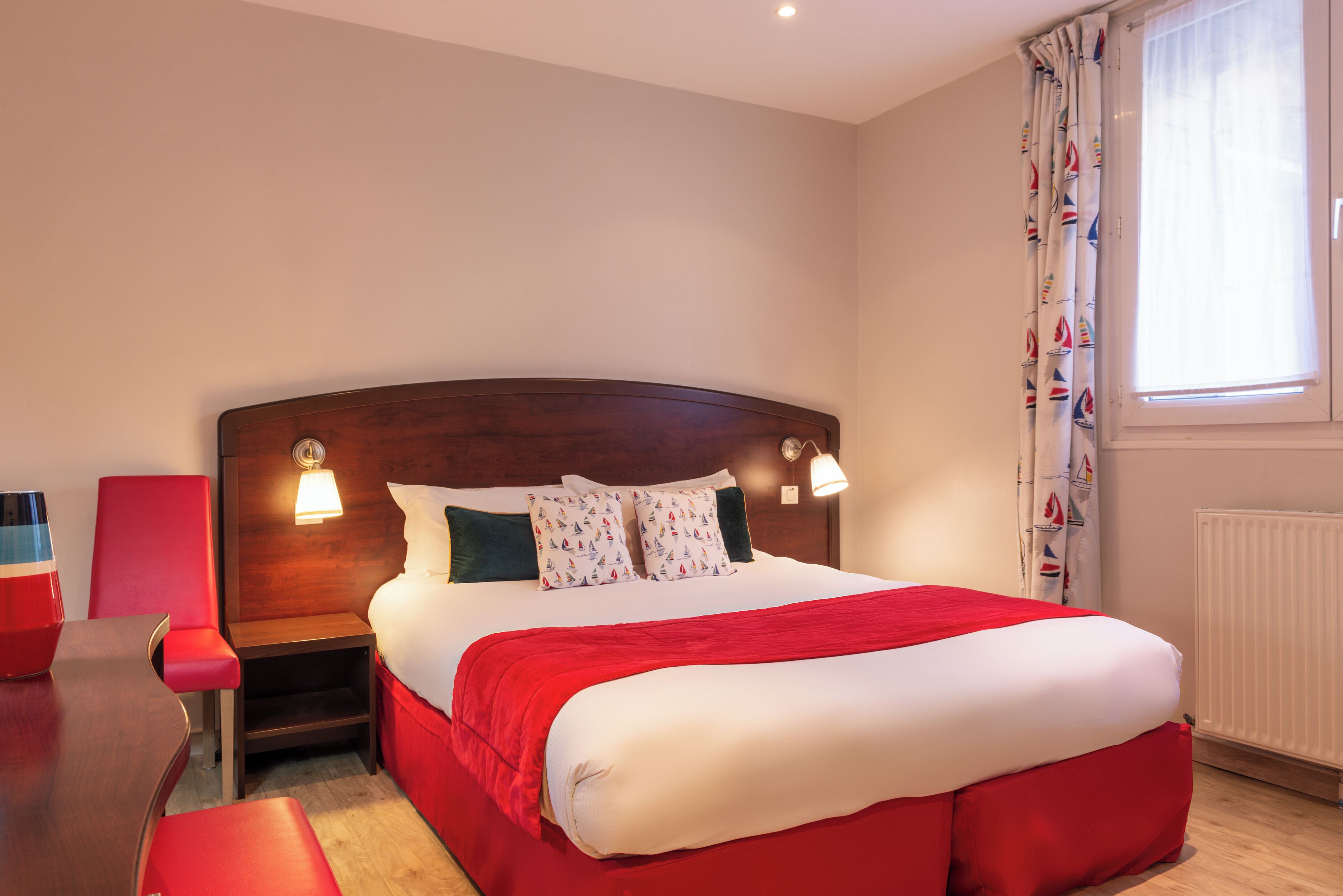 Une chambre d’hôtel avec un grand lit, une tête de lit en bois, deux lampes de chevet, un fauteuil rouge et une fenêtre avec des rideaux.