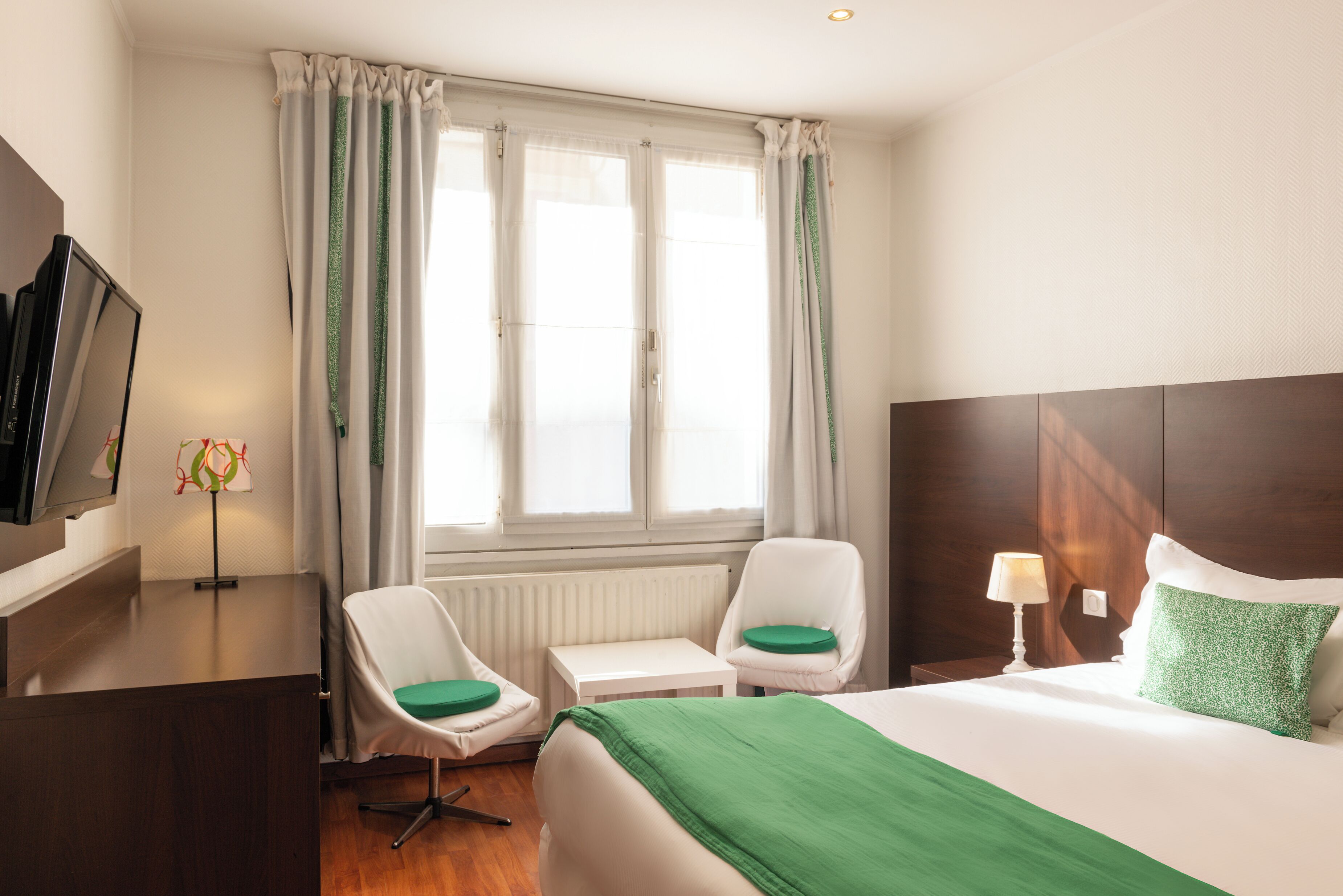 Ett hotellrum med en säng, två stolar, ett skrivbord, en tv och ett fönster med gardiner.
