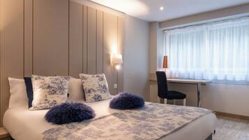 Una cama bien hecha con ropa de cama estampada y cojines decorativos, una lámpara de noche y vistas a una ventana con cortinas translúcidas.