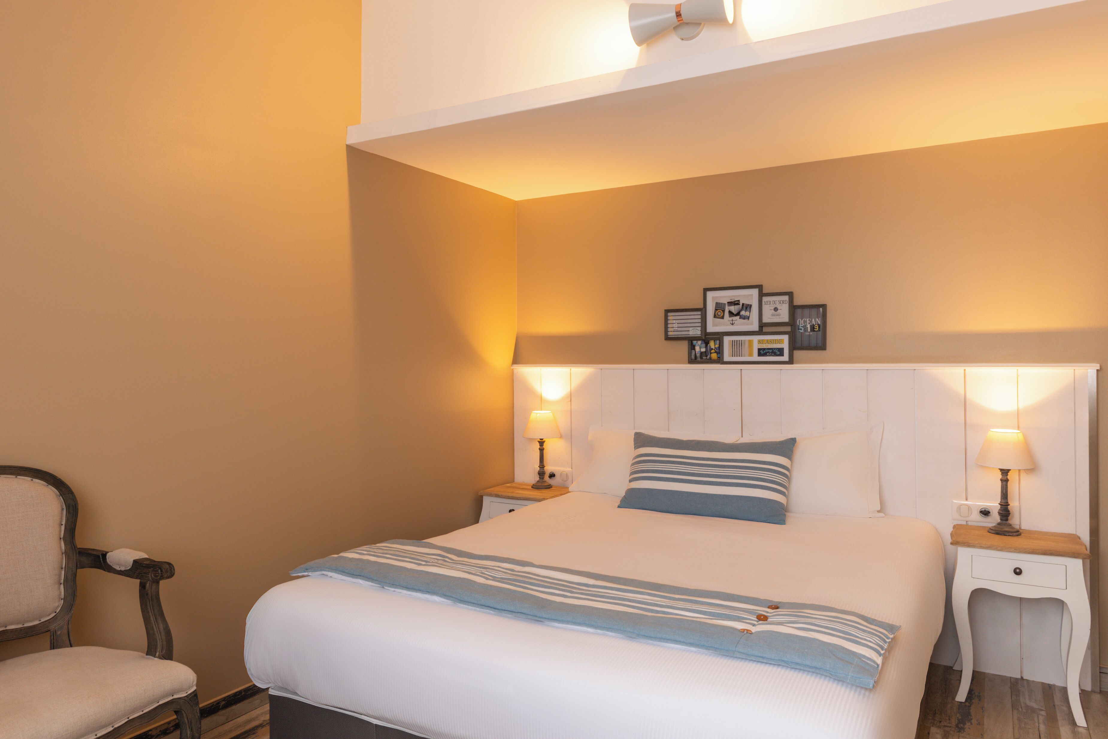 Habitación de hotel ordenada con una cama grande, dos mesitas de noche con lámparas, una silla y un cabecero con cuadros.