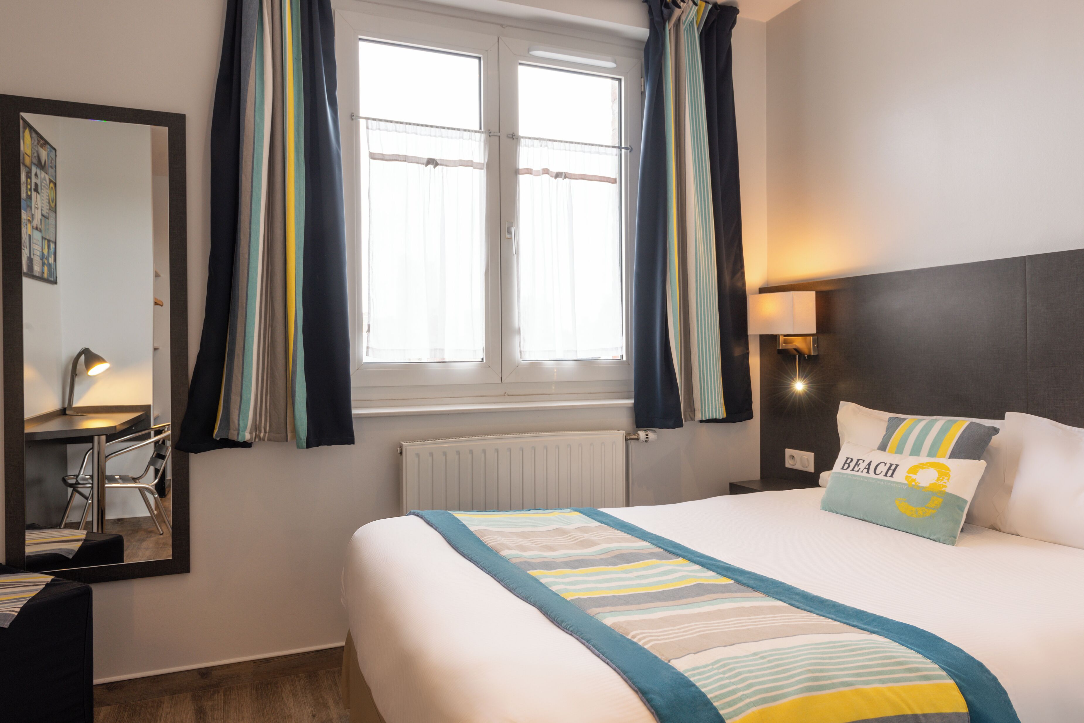 A hotel room with a large bed, a bedside table with a lamp, a mirror, and a window with curtains.