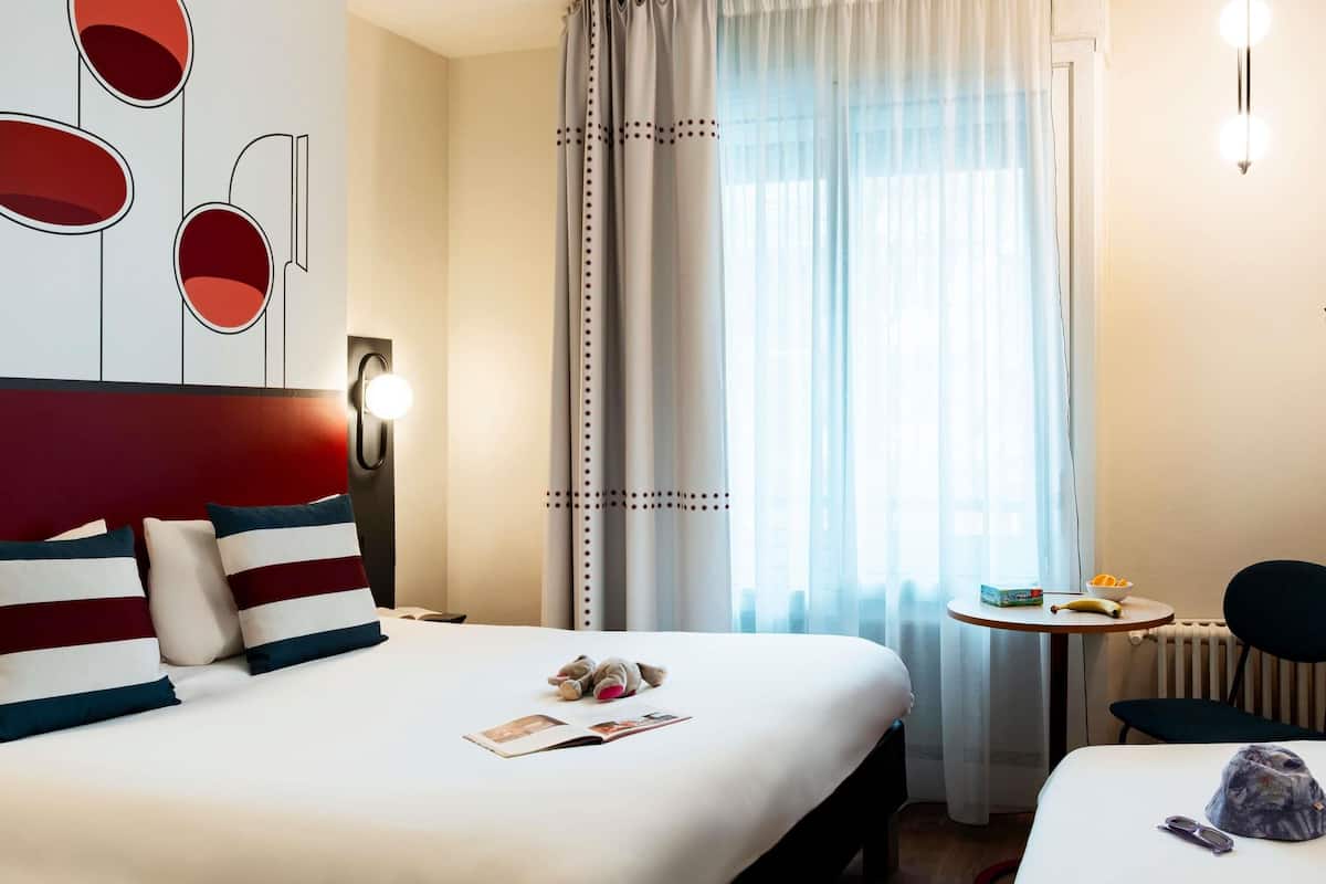 A hotel room with a large bed, a small table with a banana and a bottle, a chair, and a wall with abstract art.