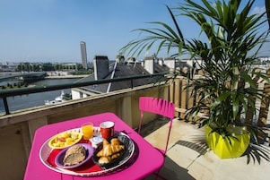 Un balcón con una mesa lista para el desayuno, una planta en maceta y vista a un río y edificios.