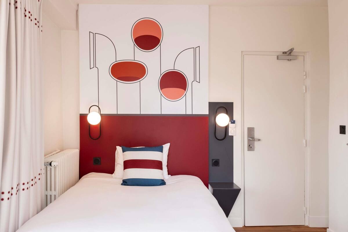 A hotel room with a bed, a red accent wall, a white radiator, a door, and a wall-mounted light fixture.