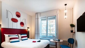 Ein modernes Hotelzimmer mit einem großen Bett, einem kleinen Tisch mit einem Laptop, einem Sessel und Blick auf die Stadt durch das Fenster.