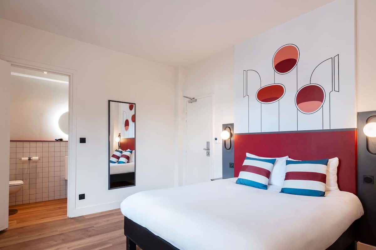 A modern hotel room with a large bed, a mirror, and abstract wall art.