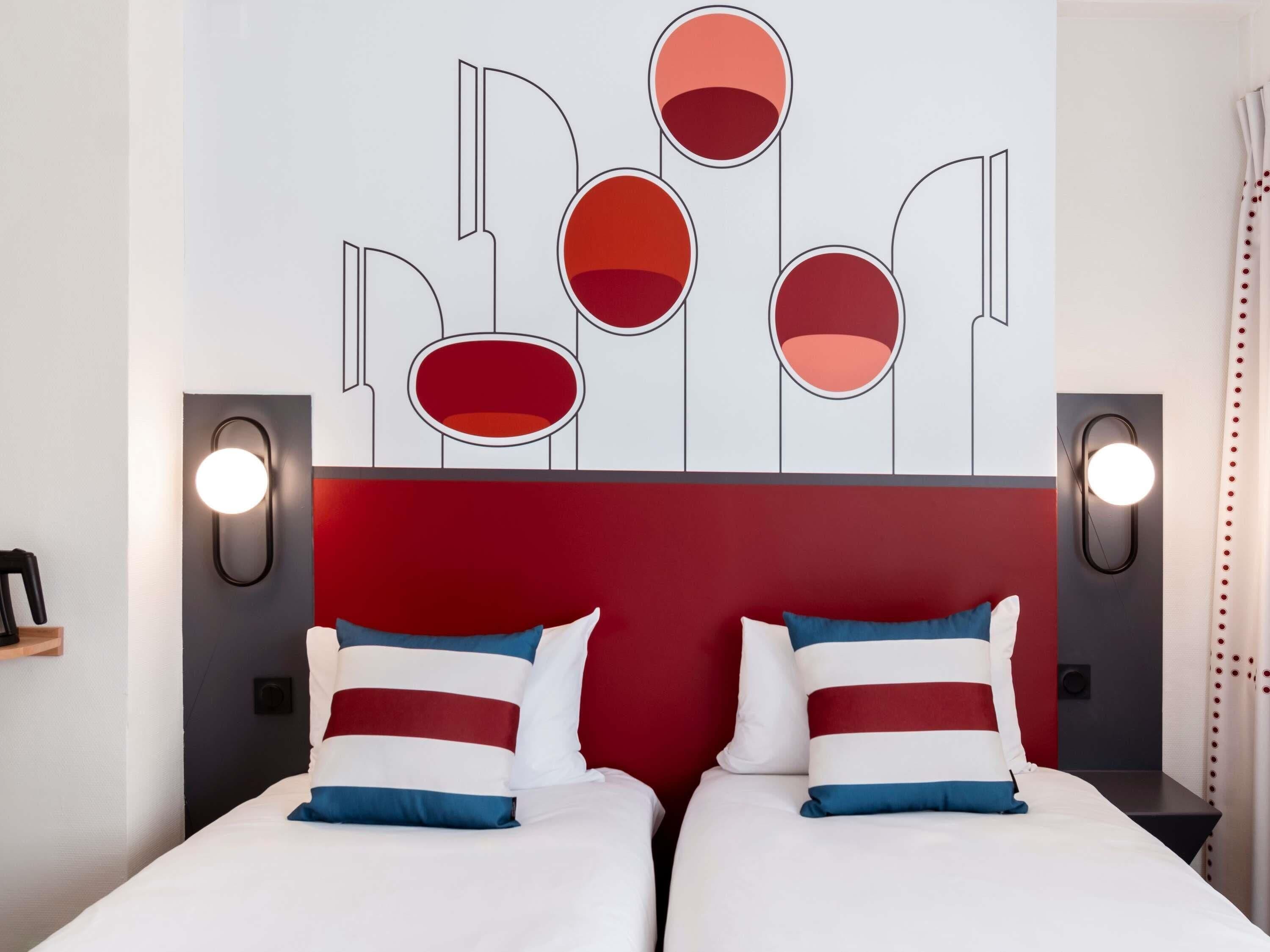 Une chambre d’hôtel dotée d’une tête de lit moderne, de deux lits avec une literie à rayures et d’appliques murales.