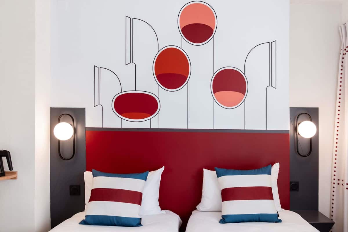Une chambre d’hôtel dotée d’une tête de lit moderne, de deux lits avec une literie à rayures et d’appliques murales.