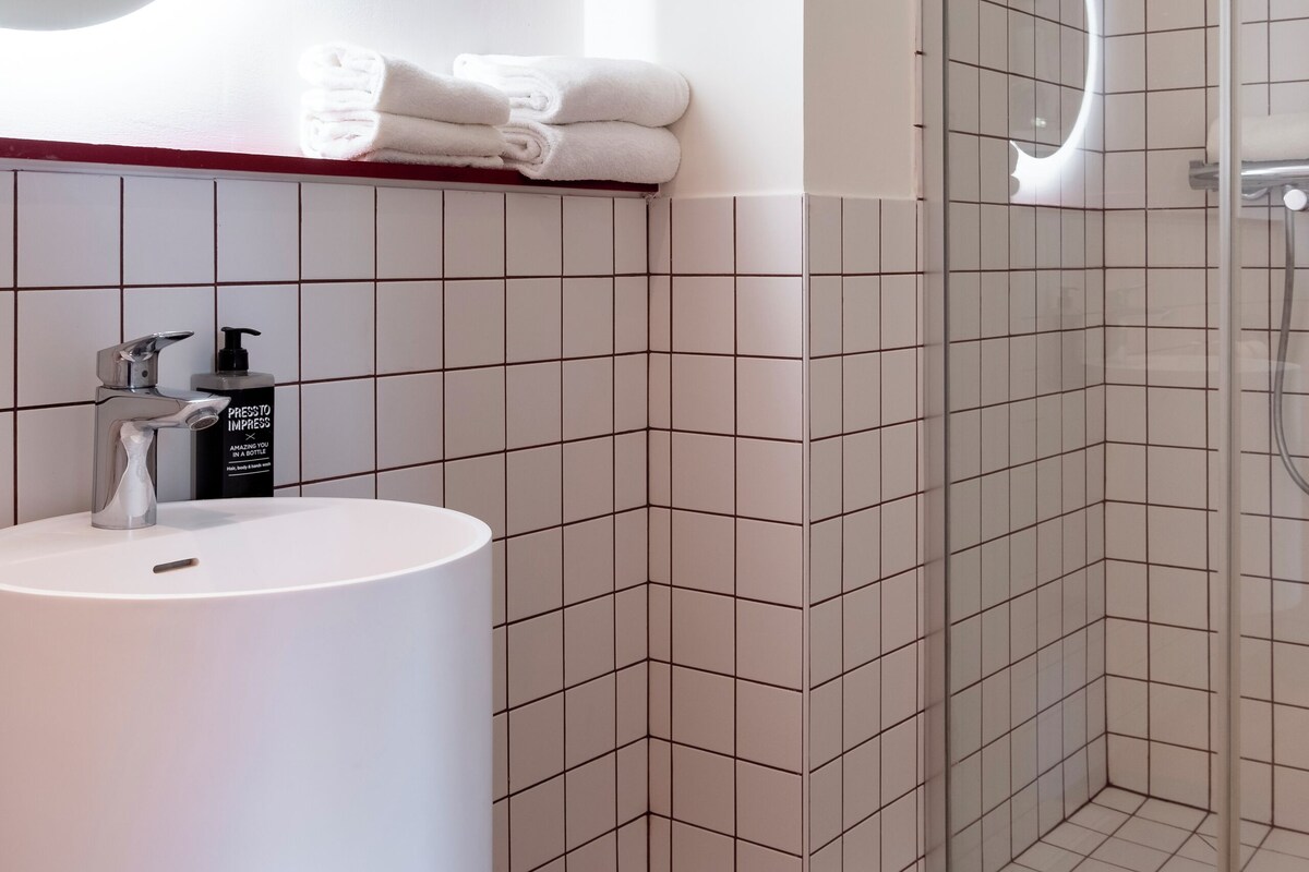 Un baño con lavamanos blanco, un espejo, ducha con mampara de vidrio y toallas ordenadas.