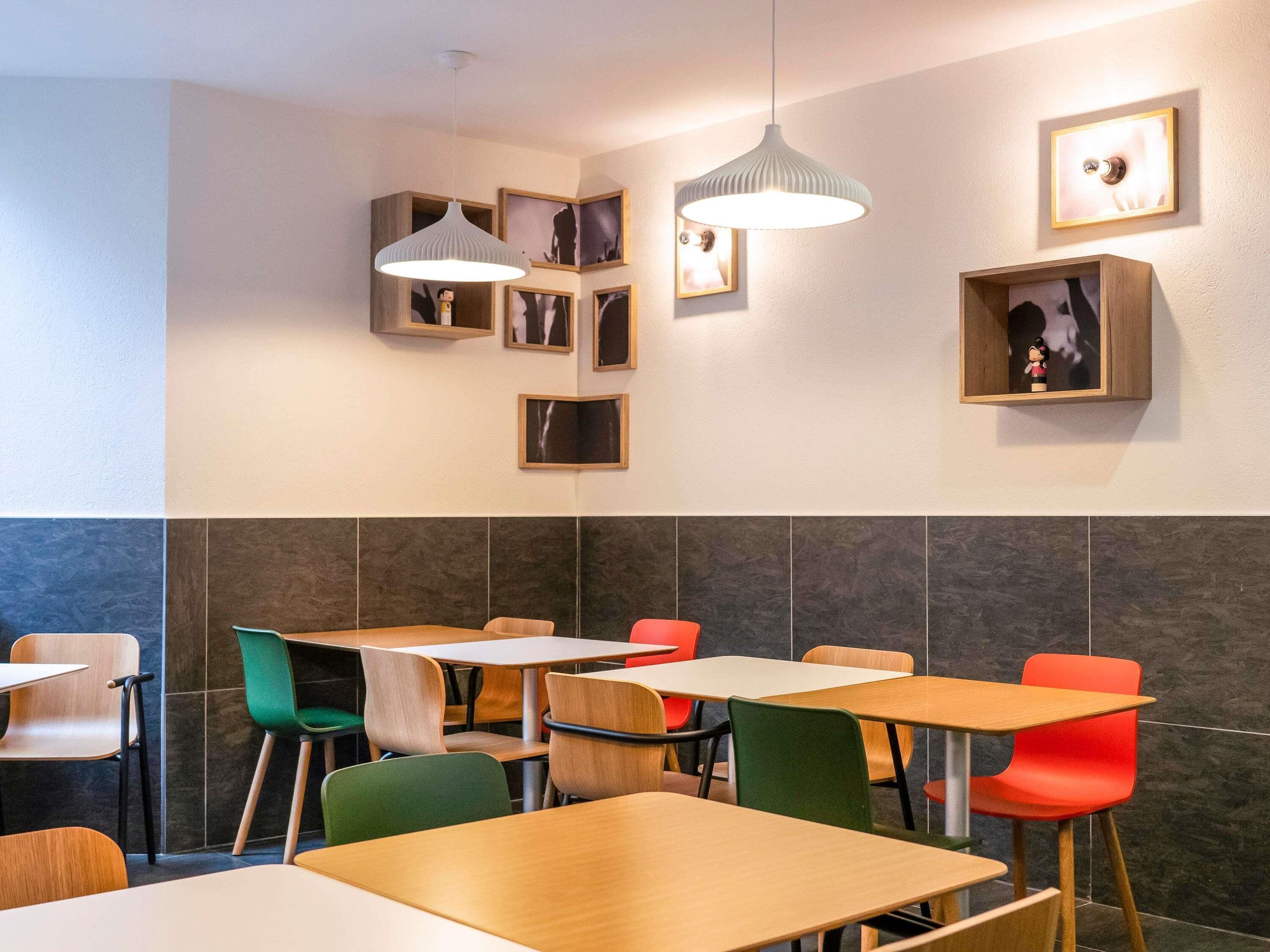 A modern dining area with wooden tables and colorful chairs, a hanging light fixture, and framed pictures on the walls.