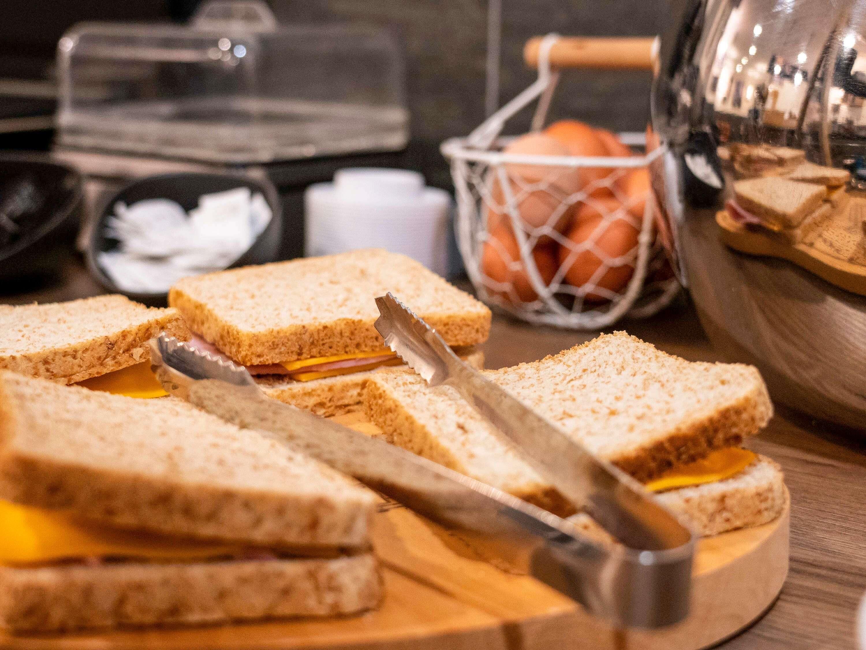 A wooden cutting board with a sandwich cut in half, a knife, and a basket of oranges in the background.