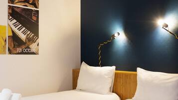 Quarto de hotel com cama, almofadas, um cartaz na parede e uma luminária.