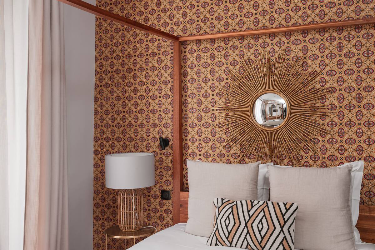 A hotel room with a patterned wallpaper, a round mirror, a bed with two pillows, a bedside table with a lamp, and a wooden headboard.